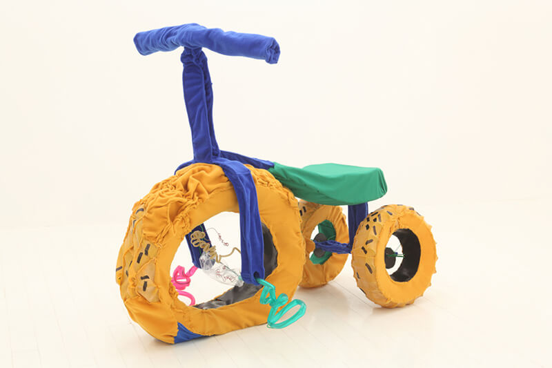 写真：松橋萌氏の立体作品。綿布やフェルト、ワイヤーで作られた三輪車の立体作品。タイヤは黄色・ハンドルは青色・サドルは緑色という鮮やかな色彩で作成されている。